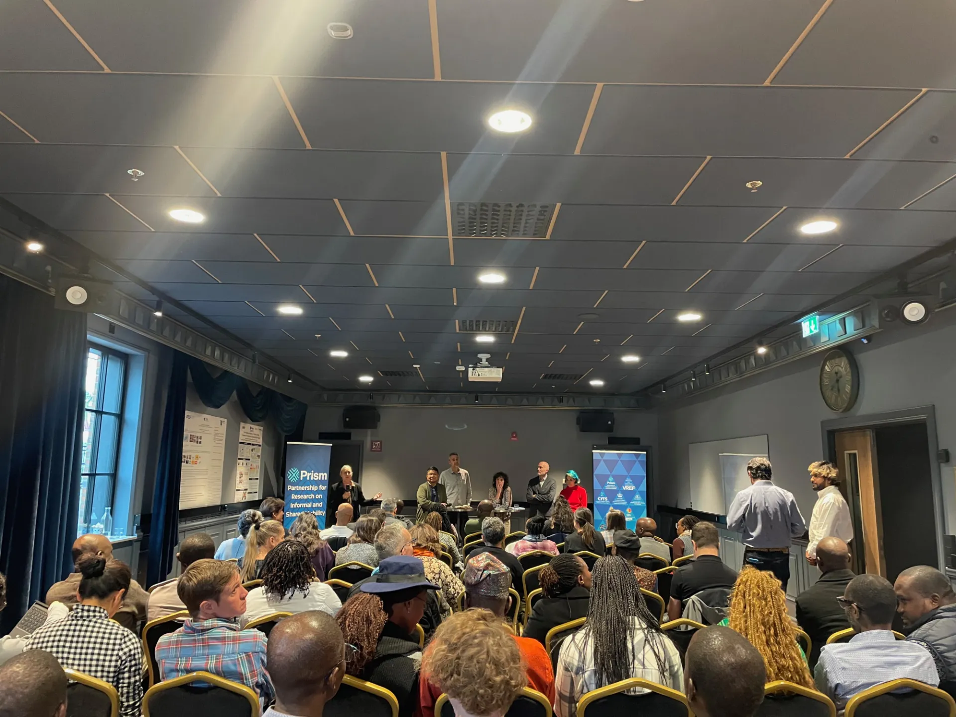 A wide-angle view of one of the sessions at the PRISM Conference in Gothenburg, showing a packed audience listening to a panel of speakers on stage. Banners for PRISM and VREF are displayed alongside the presentation screen.